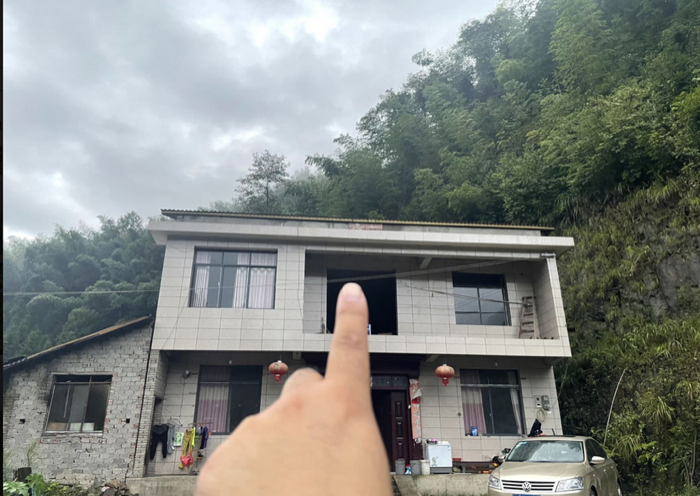 两层乡间别墅的正面，背后是长满竹林的大山；手指指向二楼中间一个没有安装窗户的空洞窗口，门口挂着红灯笼，旁边停着一辆轿车