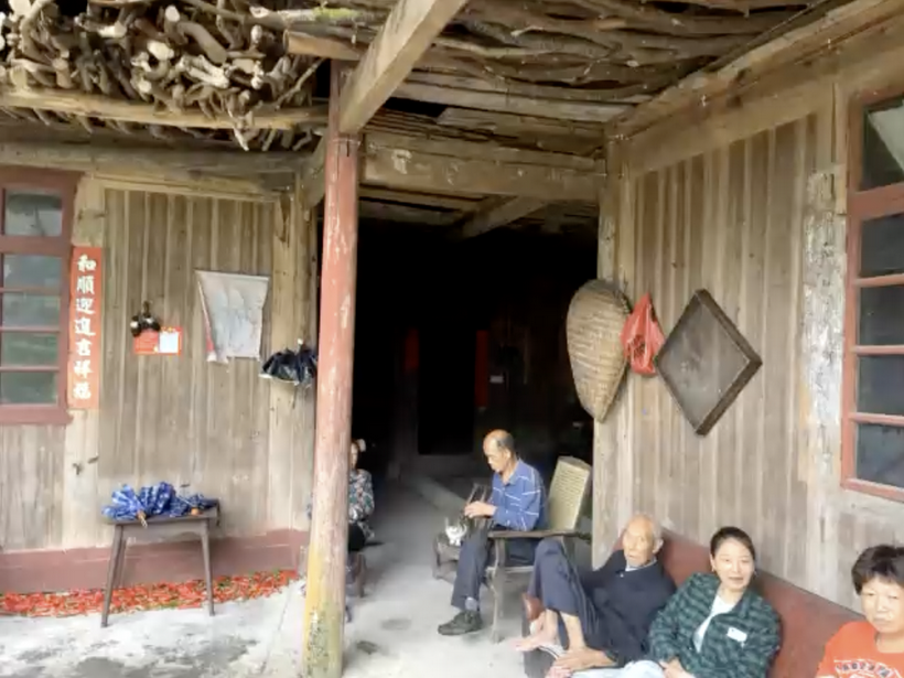 木结构老屋的门廊：木板墙上贴着红纸对联，梁上堆着柴火，墙上挂着簸箕和竹匾，几位家人坐在廊下的木凳和竹椅上，地上晾着红辣椒