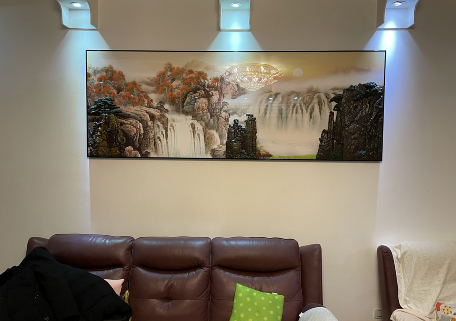 客厅深色皮沙发背后的北墙上，挂着一幅横幅很宽的山水画，画中有红叶、山石和瀑布，上方三盏射灯照着