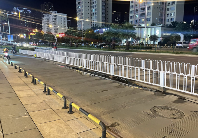 夜晚的商场外围路边：一长排白色金属栅栏沿马路延伸，人行道一侧还有黄黑隔离桩，把人行道和车道完全隔开