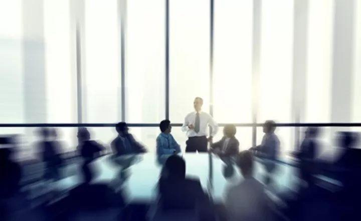 配图：明亮的会议室里，一名男子站在落地窗前讲话，围坐在长桌旁的员工身影呈动态模糊