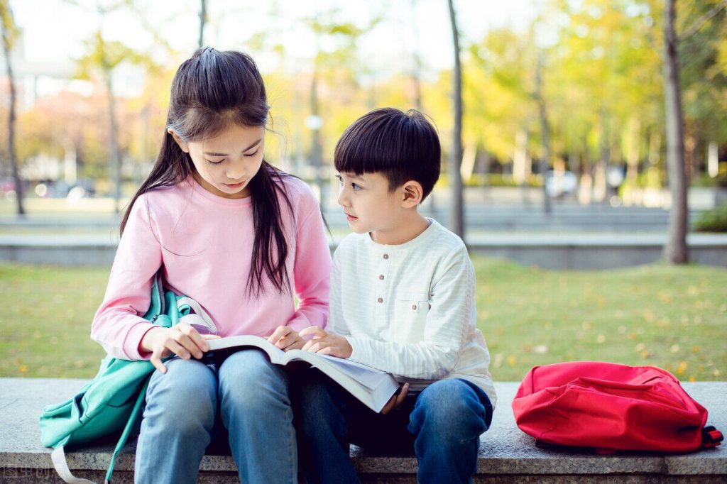 公园石凳上，穿粉色上衣的女孩和穿白色上衣的男孩一起看一本书，旁边放着红色书包