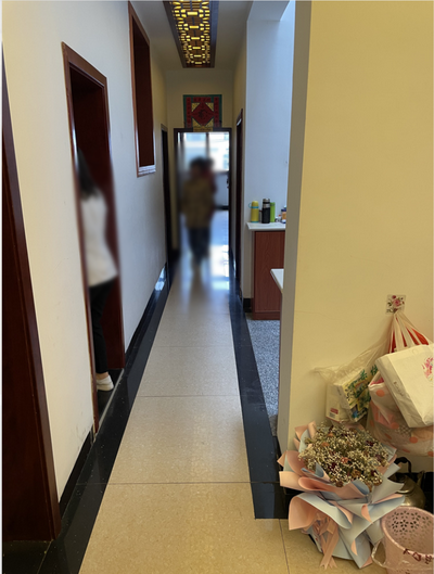 同一条长走廊的另一视角，尽头是一扇透亮的窗户，走廊里有人走动（原图已模糊处理），右侧地上堆着花束和礼品袋