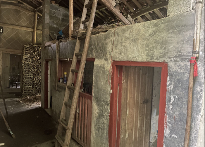 震位的猪圈：昏暗的屋内用水泥矮墙隔出几间圈栏，红色门框的木门，墙边斜靠着木梯，旁边码着成堆的柴火