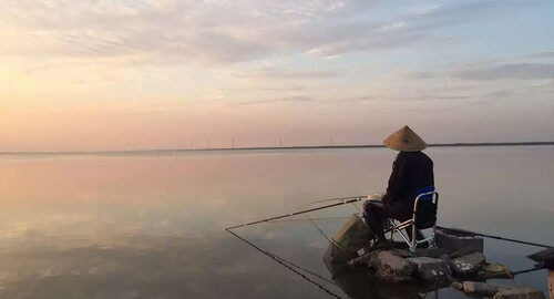 清晨平静的湖面上，一位戴斗笠的人坐在岸边石头上的折叠椅里垂钓
