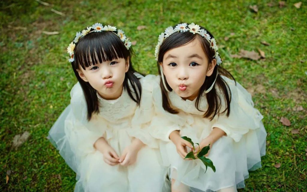配图：两个头戴雏菊花环、身穿米白色纱裙的双胞胎小女孩坐在草地上，一起嘟嘴看向镜头