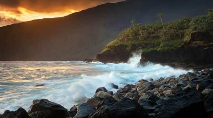 夕阳下的海岸：浪花拍打黑色礁石，远处山坡覆盖着绿色植被，天空橙黄