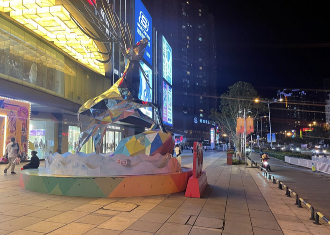 从金属鹿另一侧拍摄：鹿雕塑后方的路边，人行道与车道之间立着白色栅栏和黄黑相间的隔离桩，原本可进出的口子被挡住