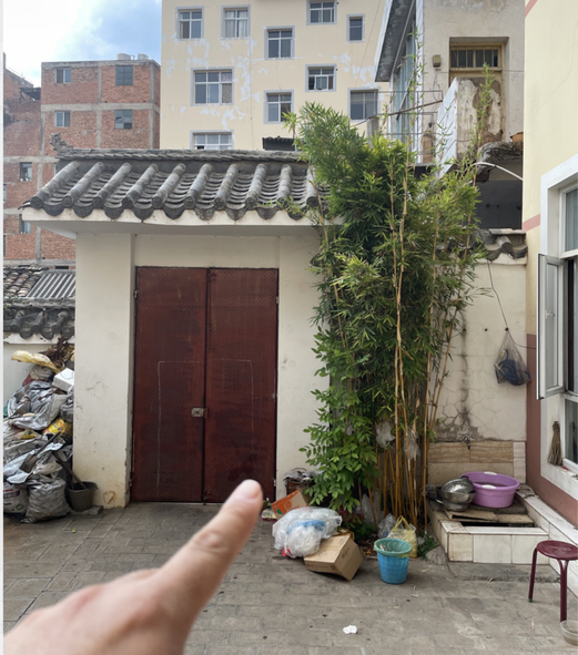 北方坎位：一间青瓦小屋开着深红色双开门，门右侧是一丛高高的竹子，再往右是摆着紫色盆子的洗手池，手指指向这扇门