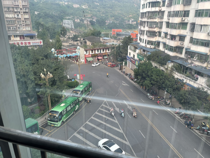 从出租屋高层窗口俯视楼下路口：几条道路在楼前交汇成夹角，车流、公交车和摩托车往来，斑马线直冲过来