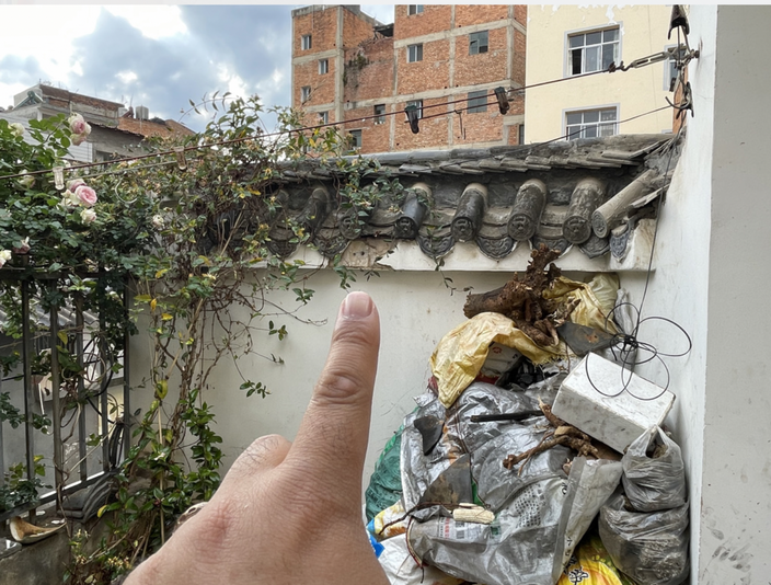 乾位围墙：手指指向墙头的青瓦装饰，瓦檐上方可见破损，下方仍是那堆袋装垃圾