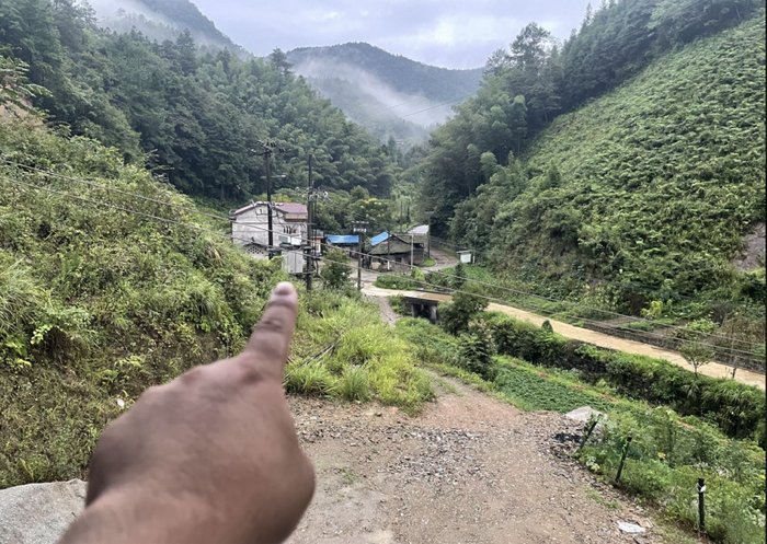 从别墅门前向西望去：手指指向山谷，一条土路通往谷中的几栋房屋，两侧青山夹峙，远处云雾缭绕，视野开阔