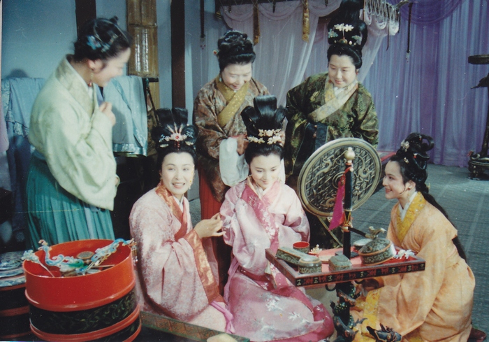 古装剧剧照：闺房中几位盛装发髻的女子围坐在一起，中间一位穿粉衣的女子对着铜镜梳妆，左下角放着红色妆奁