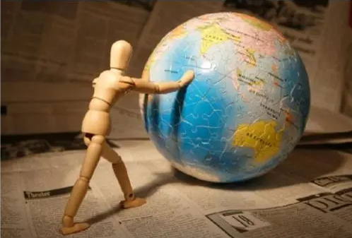 配图：木质人偶模型用双手推动一个比它还大的拼图地球仪，地面铺着报纸