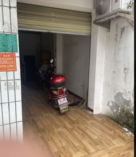 诊所店外东北侧：卷帘门半开的门口停着一辆红色电动车，右侧外墙墙皮大面积剥落、底部发霉发黑，墙上挂着空调外机